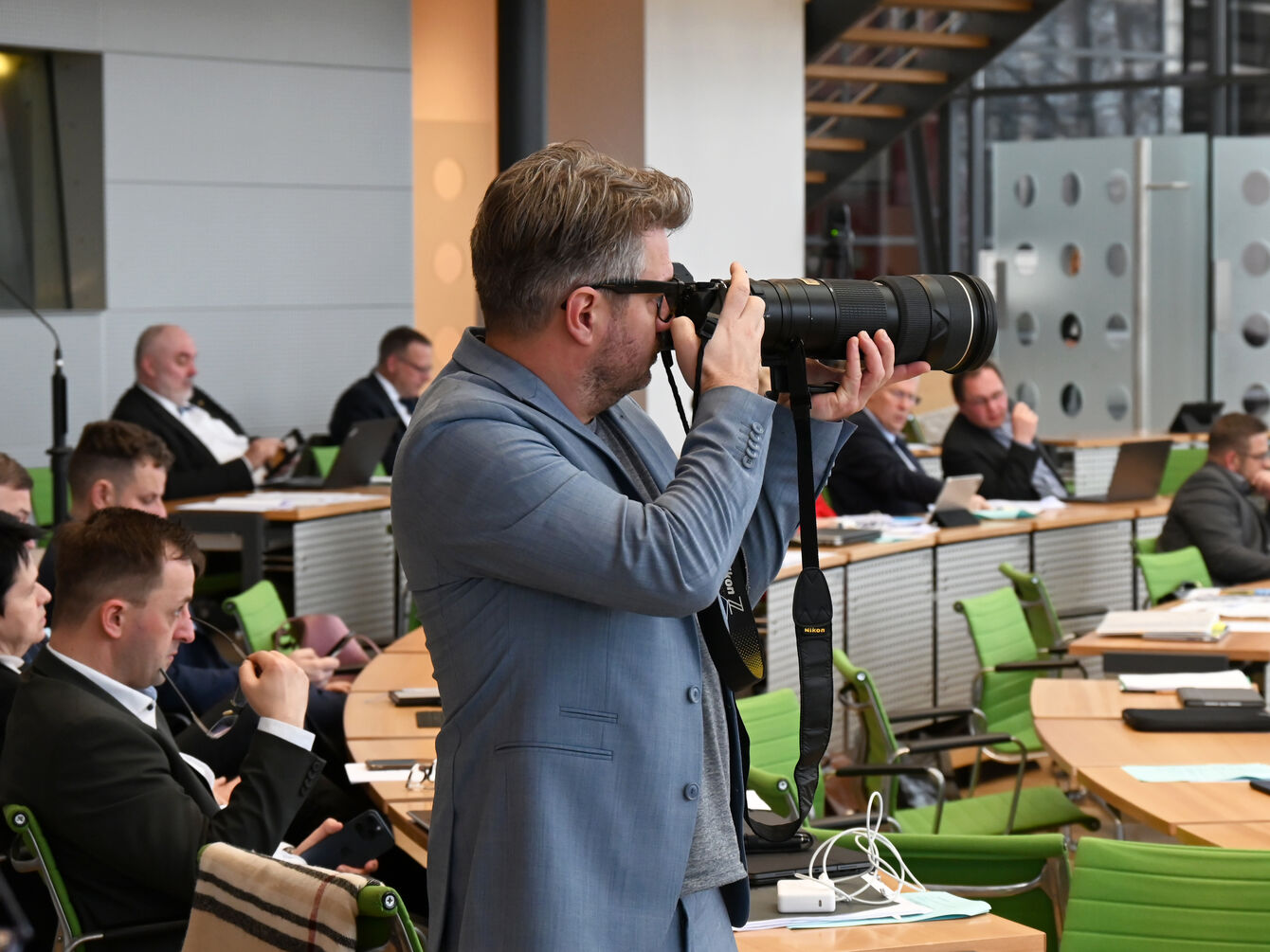 Im Plenarsaal steht zwischen den Reihen der Abgeordnetensitzplätze ein Fotograf im grauen Anzug und einer Kamera mit großem Objektiv vor dem Gesicht.