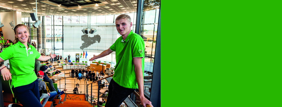 Eine junge Frau und ein junger Mann mit Landtagsshirts mit einladender Geste auf der Besuchertribüne des Landtags