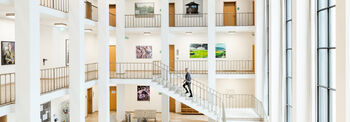 Im Altbau Foyer geht eine Frau eine Treppe hinauf. Auf den zwei oberen Gängen hängen viele Kunstwerke an den Wänden.