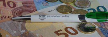 Auf mehreren Geldscheinen liegt zwischen Geldmünzen ein Kugelschreiber mit Landtags Logo.