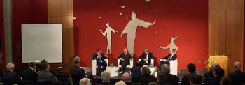 Im Ständehaus sitzen in einem großen Saal vier Diskutanten auf einem Podium vor einer roten Wand, viele Personen auf Stühlen im Publikum hören ihnen zu.