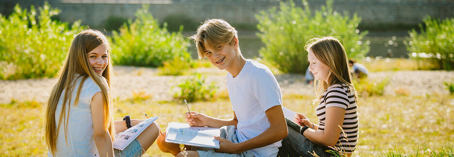 Zwei Mädchen und ein Junge sitzen mit Schulsachen auf der Elbwiese und schauen in ein Buch. Im Hintergrund ist der Landtag zu sehen.