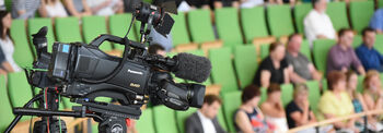 Eine Videokamera steht auf der Besuchertribüne, im Hintergrund sitzen Gäste auf Plätzen.