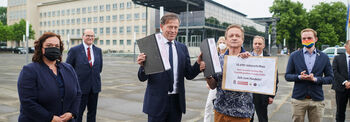 Übergabe einer Petition auf dem Landtags-Vorplatz. In der Mitte steht der Landtagspräsident mit einem Aktenordner in der rechten Hand, einen zweiten Ordner hält er in der linken Hand, gemeinsam mit einem Mann, der zusätzlich ein Schild hält. Um die beiden