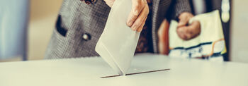 Eine Person im grauen Mantel steckt einen Stimmzettel in eine Wahlurne.