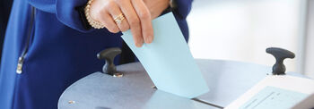 Eine Frau in blauem Mantel steckt einen hellblauen Stimmzettel in eine Wahlurne.