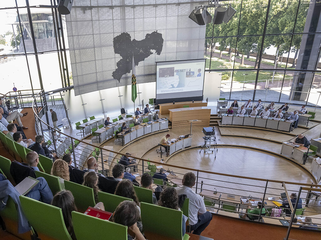 Plenarsaal während einer Anhörung mit Leinwand, Sachkundigen und Besuchern auf der Gästetribüne.