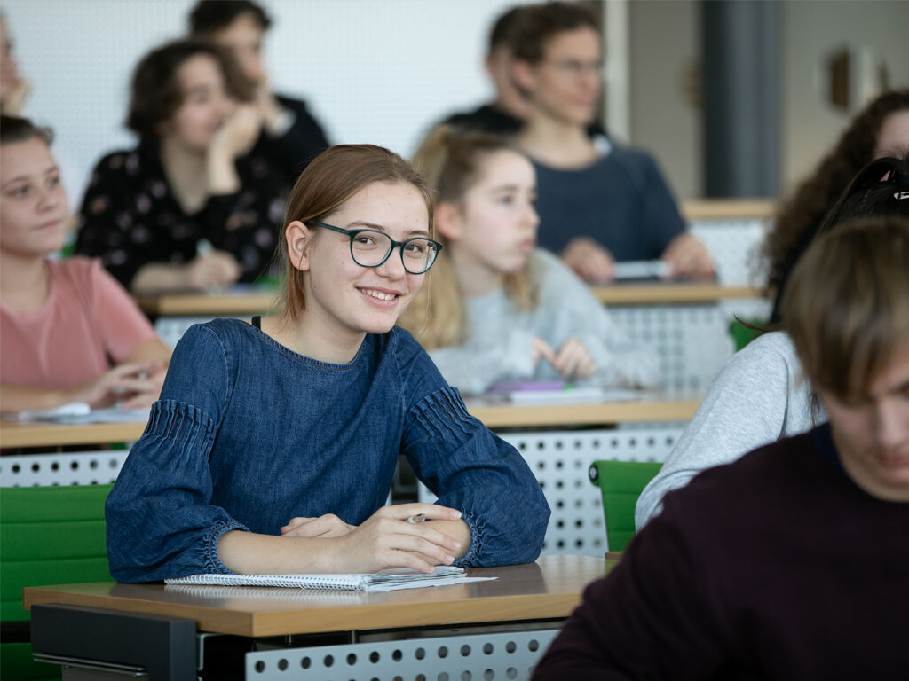 Ein junges Mädchen mit Brille sitzt im Plenarsaal, um sie herum sitzen weitere Kinder.