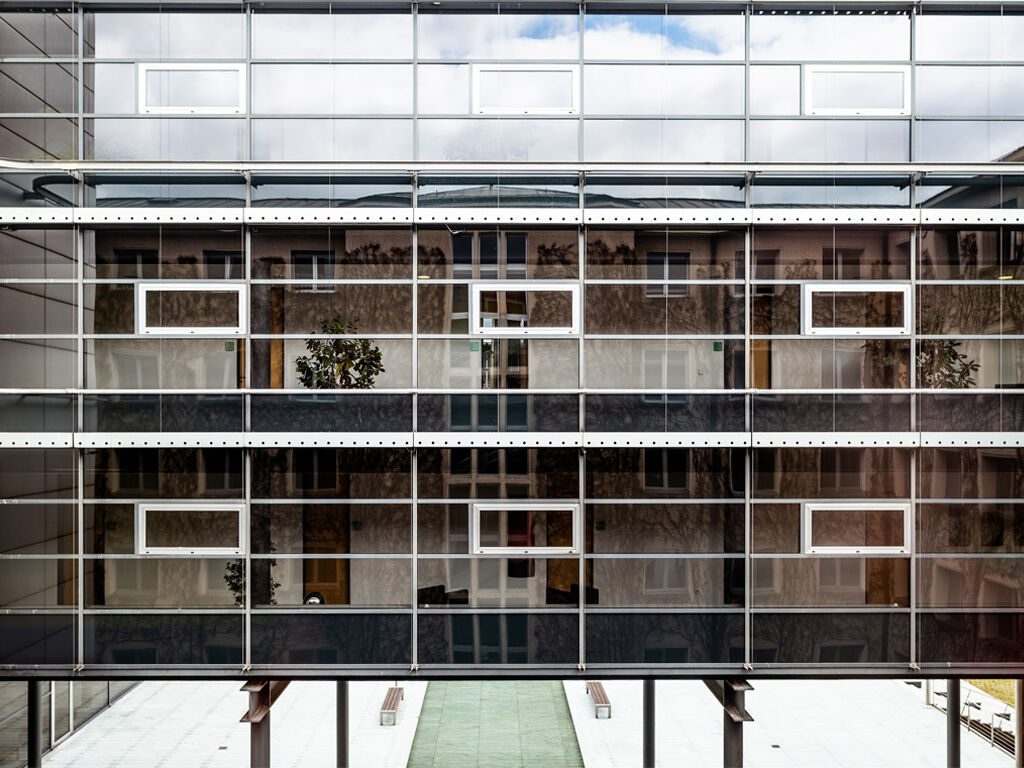 Ein verglastes Gebäude über drei Stockwerke, in den Fenstern spiegelt sich teilweise der blaue Himmel mit Wolken.