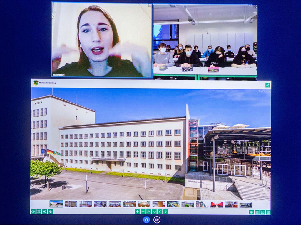 Auf einem großen Bildschirm findet eine Videokonferenz statt. Zwei Kacheln nebeneinander zeigen links die Besucherführerin und rechts eine Schulklasse in einem Klassenzimmer. Darunter in einer großen Bildkachel ist ein Screenshot abgebildet.