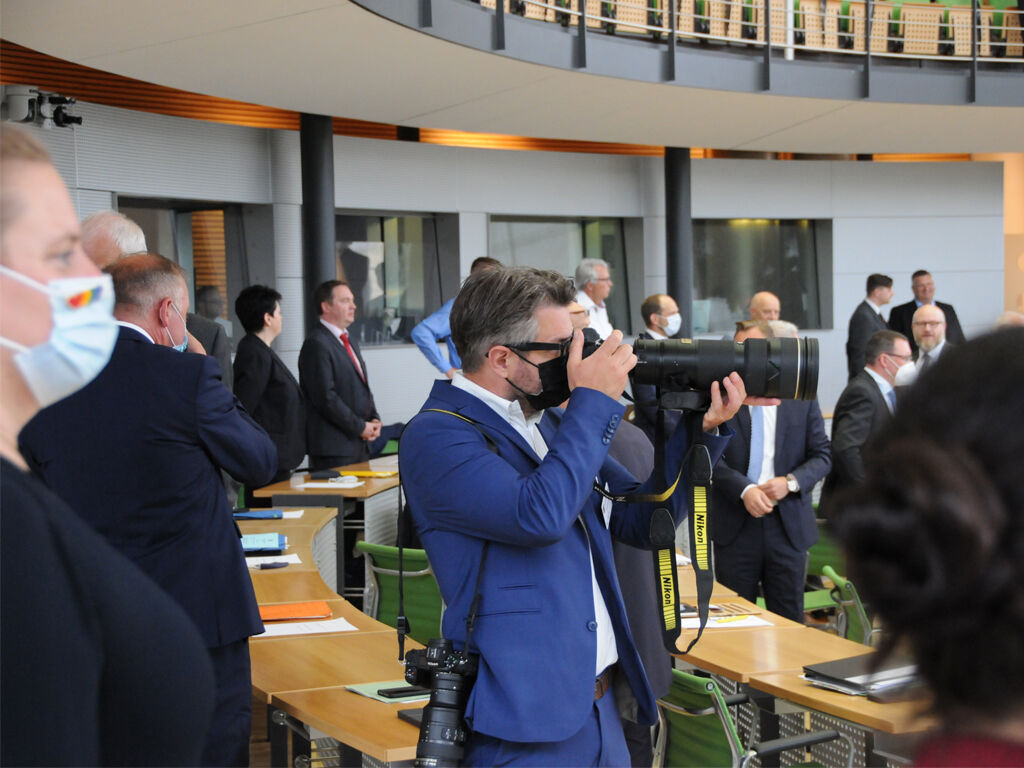 Im Plenarsaal steht zwischen den Reihen der Abgeordnetensitzplätze ein Fotograf im blauen Anzug und einer Kamera mit großem Objektiv vor dem Gesicht.