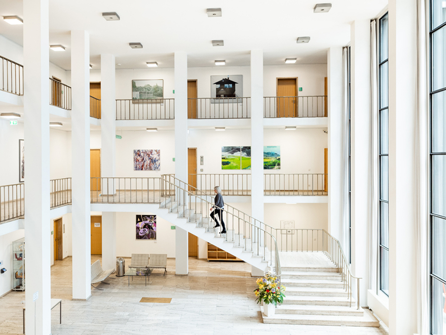 Blick in das offene Foyer des Landtag-Altbaus. Auf mehrern Etagen hängen Kunstwerke an den Wänden, vom Erdgeschoss in den ersten Stock führt eine Treppe, auf der eine Frau zu erkennen ist.