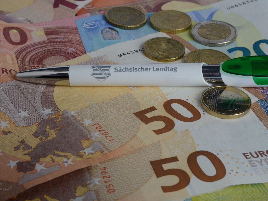 Auf mehreren Geldscheinen liegt zwischen Geldmünzen ein Kugelschreiber mit Landtags Logo.