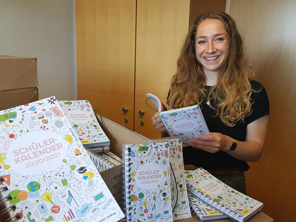 Eine junge Frau hält einen Schülerkalender in der Hand und lächelt in die Kamera. Vor ihr liegen noch mehr Schülerkalender in einem Karton.