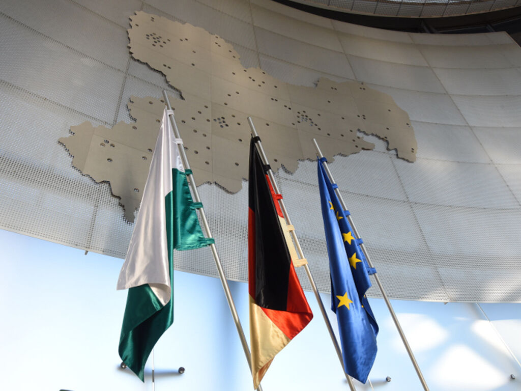 Vor einer großen silbernen Sachsenkarte aus Metall stehen drei Fahnen. Sie stehen für Sachsen, Deutschland und Europa.