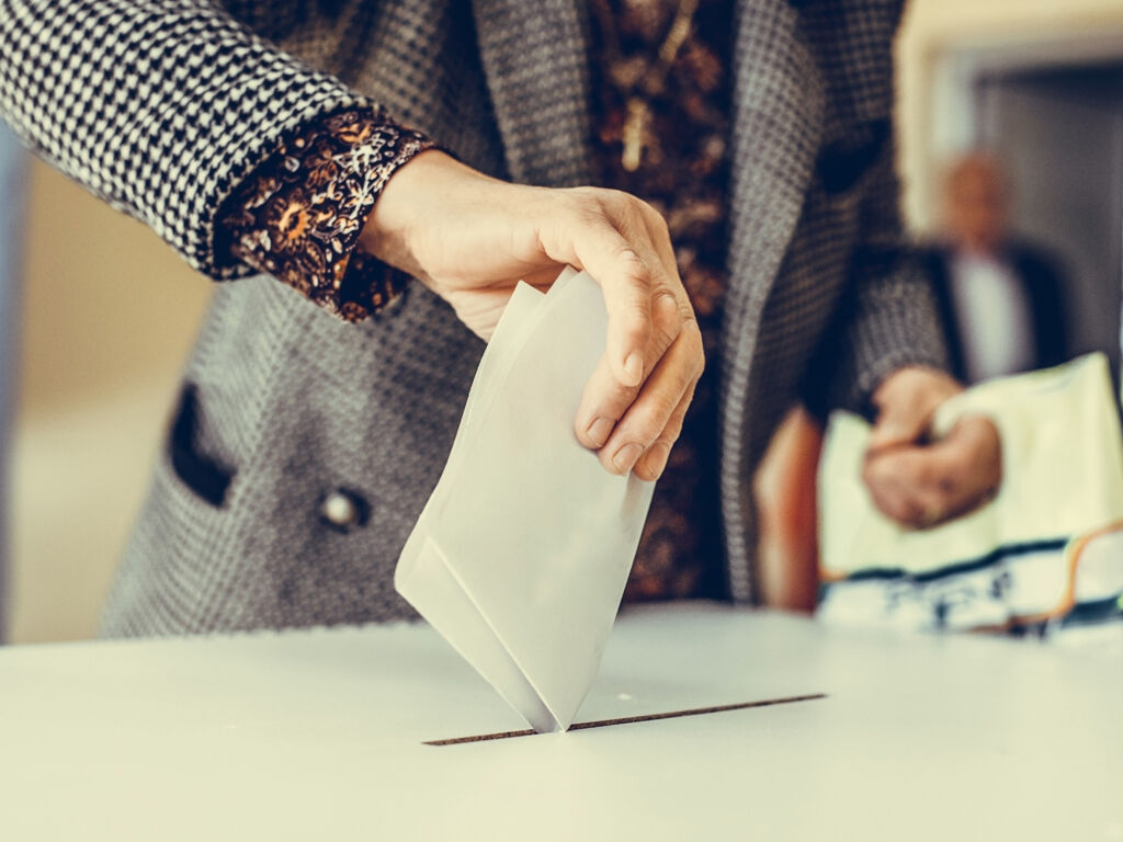 Eine Person im grauen Mantel steckt einen Stimmzettel in eine Wahlurne.
