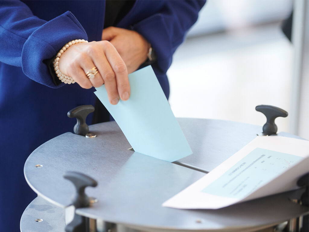  Eine Frau in blauem Mantel steckt einen hellblauen Stimmzettel in eine Wahlurne.