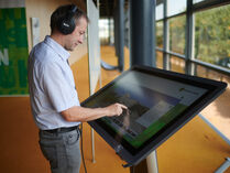 Ein Mann mit Kopfhörern steht vor einem großen Touchscreen-Bildschirm und schaut sich den Virtuellen Rundgang an.