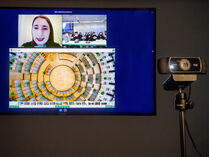 Auf einem großen Bildschirm wird eine Videokonferenz in Form einer digitalen Führung abgehalten. Im Vordergrund steht rechts außerdem eine kleine Webcam.