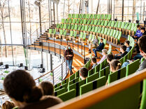 Auf der Besuchertribüne sitzen Schülerinnen und Schüler. Vor ihnen an der Brüstung steht eine Gruppenführerin.