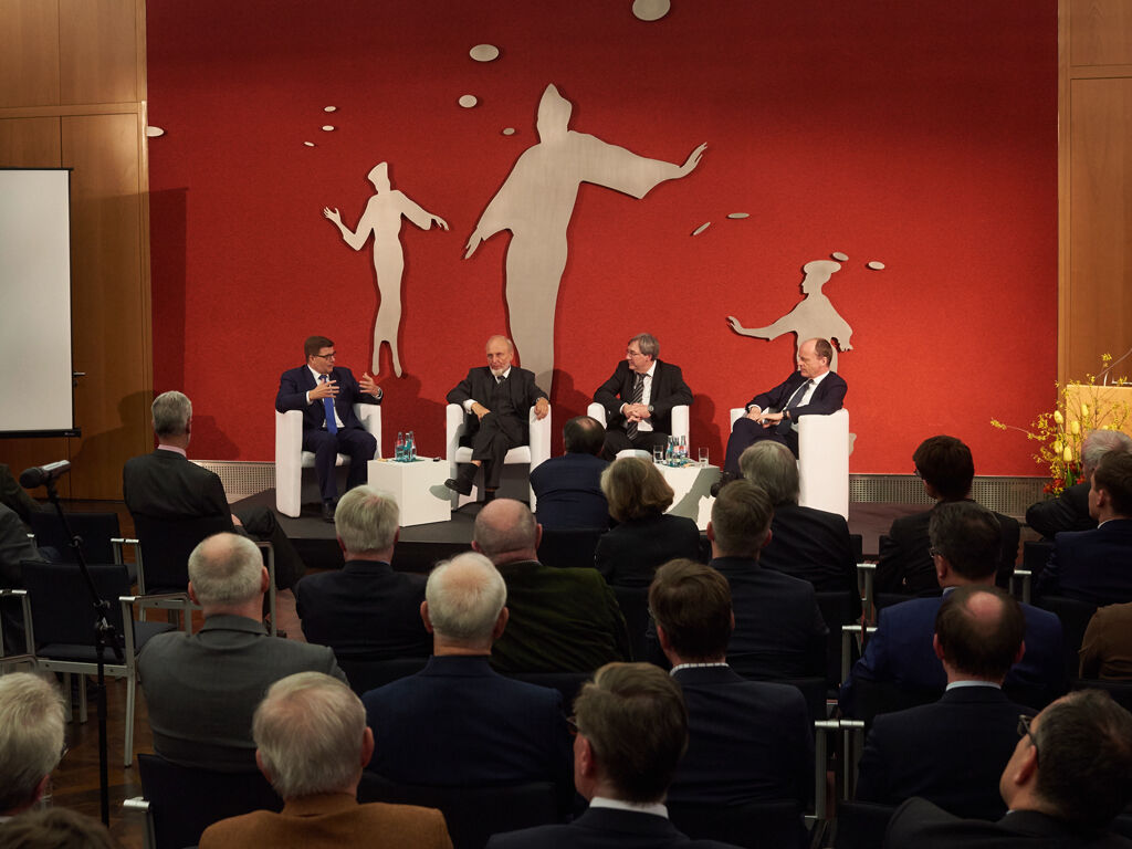 Im Ständehaus sitzen in einem großen Saal vier Diskutanten auf einem Podium vor einer roten Wand, viele Personen auf Stühlen im Publikum hören ihnen zu.