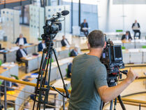 Auf der Pressetribüne des Plenarsaals stehen zwei Videokameras. Die rechte wird von einem Kameramann bedient, der das Geschehen unter ihm im Plenum filmt.