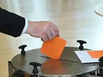 Hand mit orangefarbigem Stimmezettel an einer Urne