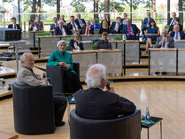 Im Rund des Plenarsaals sitzen drei Personen in Sesseln, Gäste hören ihnen beim Gespräch von ihren Sitzplätzen aus zu.