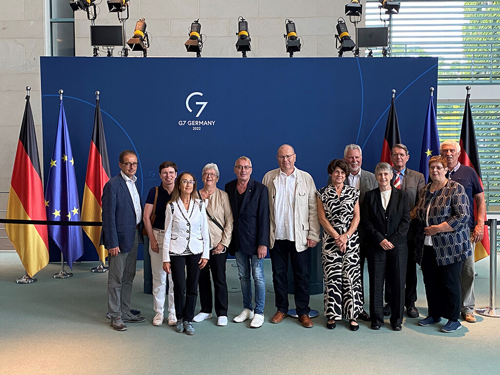 Gruppenbild der ehemaligen Abgeordneten vor einer blauen Pressewand vom G7-Gipfel, linsk sind Deutschland- und Europafahnen zu sehen.