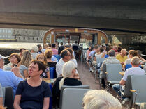 Menschen sitzen auf einem Schiff, das gleich unter einer Brücke hindurch fährt.