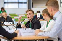 Teilnehmer am Tisch während der Vorrunde beim Argumentieren, im Hintergrund sitzt Publikum.