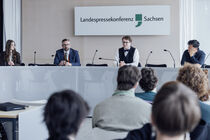 Drei Abgeordnete und eine Moderatorin sitzen im Podium der Landespressekonferenz vor der Besuchergruppe.