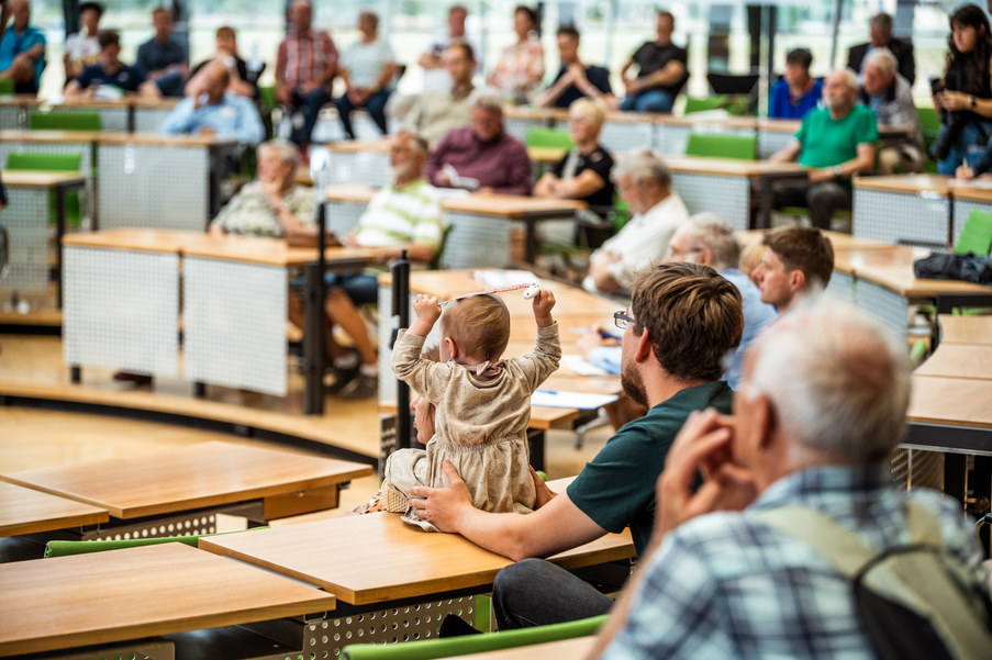 Ein Baby das auf einem Abgeordnetentisch im Plenarsaal sitzt, gehalten von seinem Vater. Im Hintergrund weitere Menschen im Plenarsaal.